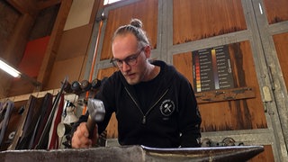 Fabian Weber hat mit einem Hammer auf ein Stück Eisen, was auf einem Amboss liegt. Im Hintergrund hängen einige Messer.