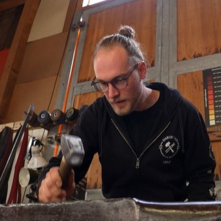 Fabian Weber hat mit einem Hammer auf ein Stück Eisen, was auf einem Amboss liegt. Im Hintergrund hängen einige Messer.