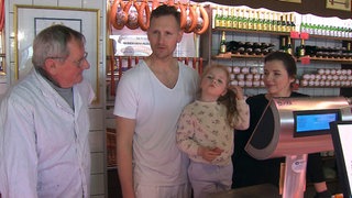 Famile Burkhardt steht in ihrer Metzgerei. Im Hintergrund hängen Würste von der Wand. Philipp hält seine Tochter mit einem Arm fest.