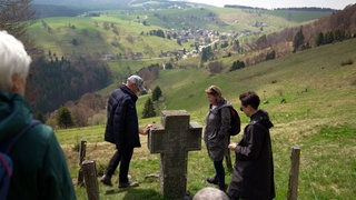 Eine Gruppe von Wanderern steht auf dem Schauinsland an einem steinernen Grabkreuz. 
