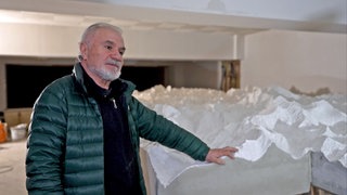 Panos Avramidis steht vor seiner weißen 3D-Nachbildung des Himalayas. Er trägt eine grüne Jacke und hält eine Hand auf der Nachbildung.