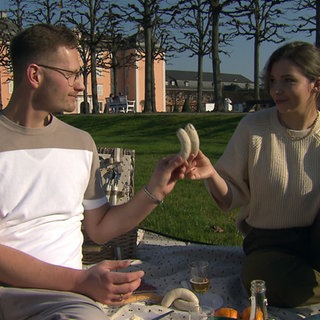 Philipp und Anna Burkhardt sitzen auf der Wiese vor dem Schloss Schwetzingen und stoßen mit einer Weißwurst in der Hand an.