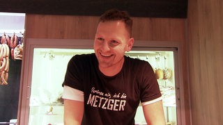Philipp Burkhardt steht in seinem Marktwagen und trägt ein schwarzes T-Shirt. Er lächelt. Im Hintergrund hängen Würste.