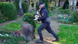 Klaus Stolzenberger steht mit seinem Prüfgerät auf einem Friedhof und lehnt sich gegen einen grauen Grabstein.