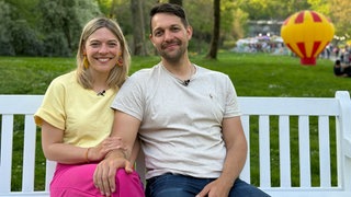 Janina und Bastian sitzen auf einer Bank und lächeln in die Kamera. Im Hintergrund ist ein rot-gelber Ballon zu sehen.