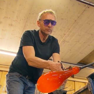 Stefan Michaelis trägt ein schwarzes T-Shirt und violett schimmernde Schutzbrille. Mit einer Zange hält er mit großem Abstand eine orange glühende Glasbirne fest.