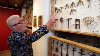 Bernhard Maurer trägt ein buntes Hemd zeigt mit dem rechten Arm auf eine Sammlung von verschiedenen Korkenziehern in seinem Museum in Burkheim.