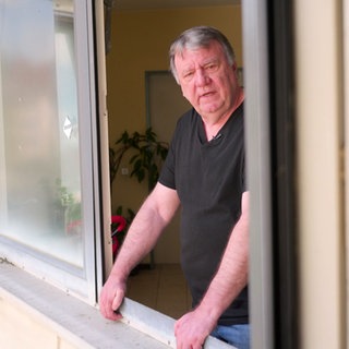 Dieter Scheibel steht an einem offenen Fenster und hält sich mit beiden Händen an der Fensterfassung fest. Er schaut Richtung Kamera. Die Fassade des Hauses ist leicht gelblich.