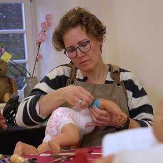 Anja Müller sitzt in ihrer Puppenklinik und wickelt einen blauen Verband um eine Puppe. Im Vordergrund sieht man verschwommen einen Puppenkopf.