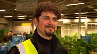 Christian Polder steht mit einer gelben Warnweste vor einer hell ausgeleuchteten Lagerhalle. Er hat lockige, braune Haare und einen Goatee. Er lächelt in die Kamera.