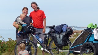 Dina und Rafael Streib stehen hinter einem schwarzen Fahrrad mit einem Kinderwagen-Anhänger. Auf ihrem Arm und vor dem Fahrrad sind zwei ihrer Kinder. Im Hintergrund erkennt man eine Küste.