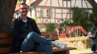 Patrik Lehmann sitzt auf einer Parkbank in einem Dorf, die im Schatten eines Baumes steht. Er lächelt in die Kamera.