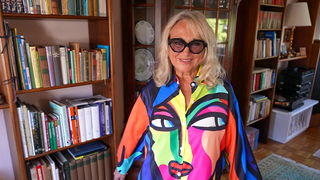Sylvia ist 77 Jahre alt. Sie steht in ihrem Wohnzimmer und trägt eine auffällige, bunte Bluse mit einem Gesicht als Motiv. Außerdem trägt sie eine große, auffällige Sonnenbrille. 