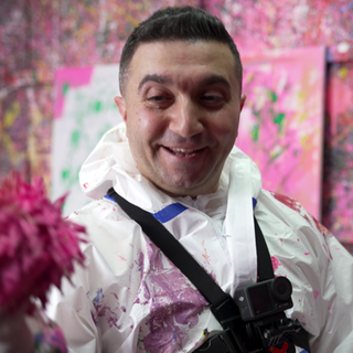 Ein Mann steht in einem Raum, in dem alle Wände voll sind von Farbspritzern. Der Mann trägt einen Schutzanzug und hält einen mit Farbe getränkten Ball in der Hand. 