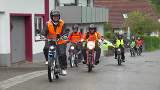 Sechs Menschen fahren auf Mofas eine Straße hoch. 