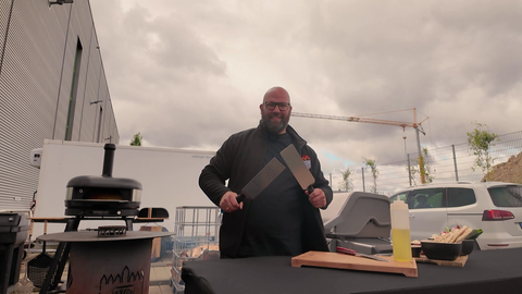 Grillmeister Ralf Ahrens steht am Grill und hält zwei Pfannenwender in der Hand.