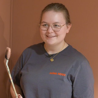 Katharina Bauer steht in einem schwarzen T-Shirt vor eine rötlichen Wand und hält einen hölzernen Pinsel in der Hand. Sie lächelt in die Kamera.