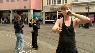Max steht in einem Tanktop in einer Fußgängerzone und hält ein Mikrofon an seinen Mund. Links von ihm tanzen zwei junge Frauen im Hintergrund.