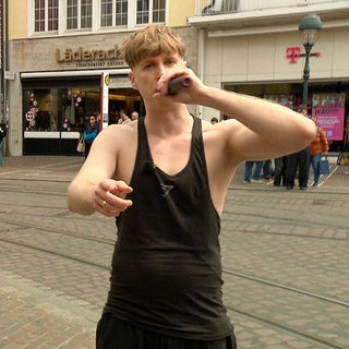 Max steht in einem Tanktop in einer Fußgängerzone und hält ein Mikrofon an seinen Mund. Links von ihm tanzen zwei junge Frauen im Hintergrund.