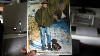 Eine Hand hält ein Bild von Georg und seinem Rauhaardackel Camillo. Gerog lächelt, Camillo steht mit seinen kurzen Beinen im Schnee.