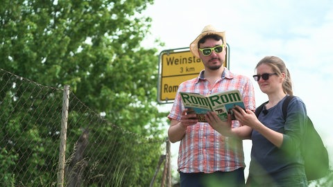 Florian Ladenburger und Laura Bieber stehen mit einem Rom-Reiseführer vor dem Ortsschild von Rommelshausen Richtung Weistadt.