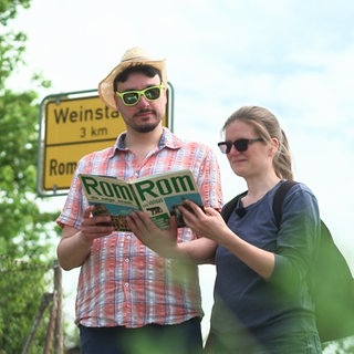 Florian Ladenburger und Laura Bieber stehen mit einem Rom-Reiseführer vor dem Ortsschild von Rommelshausen Richtung Weistadt.