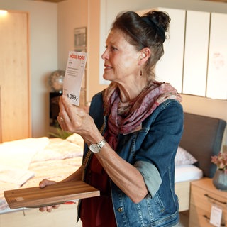 Verkäuferin Annette Schramm steht in einer Schlafzimmer-Nachbildung im Möbelhaus und hät ein Preisschild in die Luft. In der anderen Hand hält sie ein Klemmbrett.