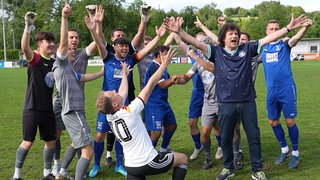 Olaf der Flipper steht mit einem dunklen Deutschland-Fußballtrikot zusammen mit Fußballern des VfB Bretten auf einem Fußballplatz. Oliver Pocher kniet vor der Mannschaft. Alle jubeln.