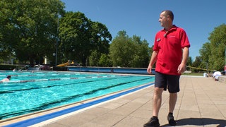 Alessandro Bagnoli trägt ein rotes T-Shirt und eine kurze schwarze Badehose. Er läuft auf der rechten Seite am Rand eines blauen Schwimmbeckens entlang.