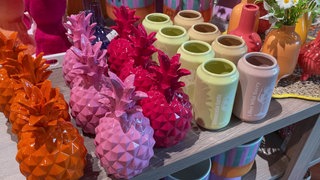 Mehrere orangene, pinke und lilane Ananas-Vasen stehen auf einem Tisch. Rechts daneben stehen schlichte Vasen in verschiedenen Farben.