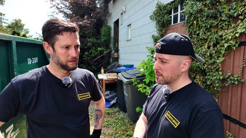 Zwei Männer, die als Entrümpler arbeiten, stehen vor einem alten Haus, neben dem ein Container steht.