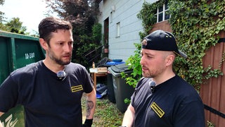 Zwei Männer, die als Entrümpler arbeiten, stehen vor einem alten Haus, neben dem ein Container steht.