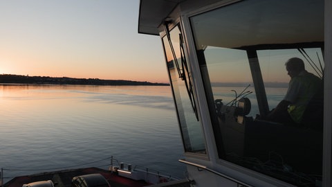Eine Fähre steht auf dem Bodensee, auf der linken Seite sieht man den Sonnenaufgang.