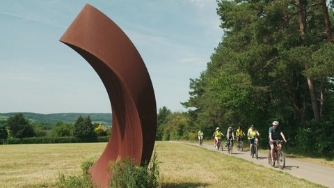 Fahrradfahrer fahren auf einem Radweg. Links im Bild ist eine Kunstskulptur zu sehen.