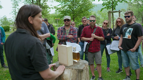 Mehrere Männer und Frauen stehen um einen Holzstamm, auf dem eine Whiskyflasche steht. 