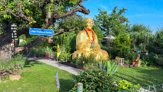Ein gepflegter Garten mit viel thailändischer Deko. Auf einem Schild steht: „Little-Thailand-Garden“. In der Mitte des Gartens ist eine 3 Meter große Buddha Statue. 