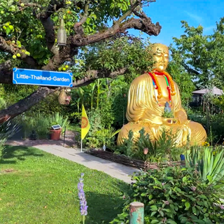 Ein gepflegter Garten mit viel thailändischer Deko. Auf einem Schild steht: „Little-Thailand-Garden“. In der Mitte des Gartens ist eine 3 Meter große Buddha Statue. 