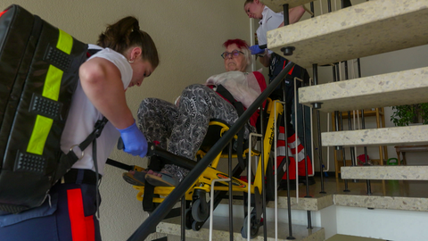 Die zwei Notfallsanitäterinnen Alina Peschers und Stephanie Dahlem tragen eine ältere Dame in einem Rettungsstuhl eine Treppe runter.