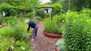 Martina Kunath steht in ihrem Kräutergarten und harkt ein Feld. Um sie herum sind verschiedene grüne Kräuterbeete mit Infokarten angelegt.
