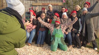 Bauer Heini steht mit seinem Team vor einem Holzgebäude. Alle tragen Weihnachtsmützen und freuen sich. Im Vordergrund macht eine Person mit einem Handy ein Foto.