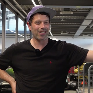Michael Höll steht im Kofferlager des Flughafens Baden-BadenKarlsruhe und stützt sich auf einen Stapel Reisekoffer ab. Er trägt eine lila Mütze und ein dunkles T-Shirt.