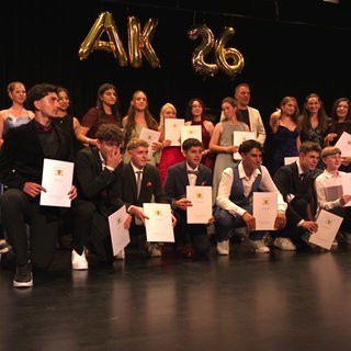 Die Abschlussklasse der Oskar-Paret-Schule steht auf einer Bühne, alle gekleidet in Anzügen oder Kleidern. Sie halten Zeugnismappen in der Hand. Im Hintegrund steht "AK26" in Ballons.