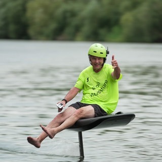 Ein Mann sitzt auf einem E-Board und zeigt mit seiner Hand den Daumen hoch. Er schwebt etwa 40 Zentimeter über der Wasseroberfläche. Er trägt einen grünen Helm. 