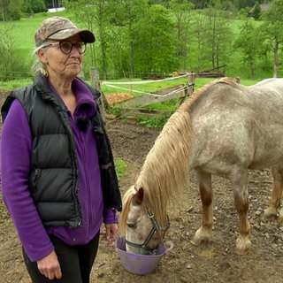 Eine Frau steht vor einem Pferd. Das Pferd frisst aus einem Trog. 