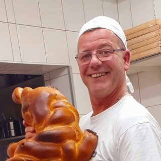 Ein Mann in Bäckerkleidung hält einen Hefezopf. Er lächelt.