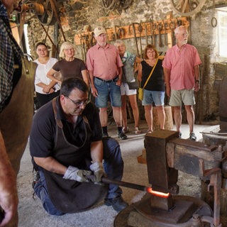 Ein Mann sitzt an einem großen Schmiedehammer. Er ist Teil einer Führung. Andere Teinehmer stehen dahinter und schauen zu.