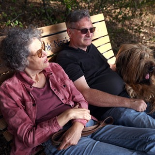 Inga und Claus Berg sitzen mit ihrem Hund auf einer Bank im Wald.