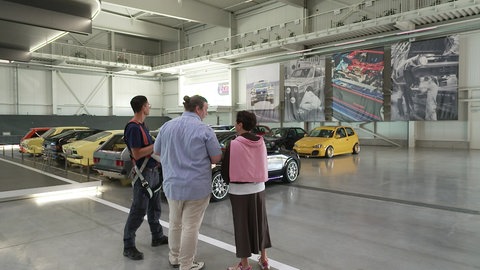 Sonja Faber-Schrecklein steht mit zwei Mitarbeitern des Technikmuseums Sinsheim in einer Autohalle. Links von ihnen stehen Oldtimer.