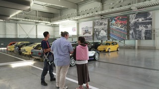 Sonja Faber-Schrecklein steht mit zwei Mitarbeitern des Technikmuseums Sinsheim in einer Autohalle. Links von ihnen stehen Oldtimer.