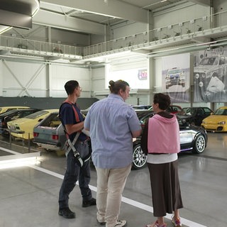 Sonja Faber-Schrecklein steht mit zwei Mitarbeitern des Technikmuseums Sinsheim in einer Autohalle. Links von ihnen stehen Oldtimer.